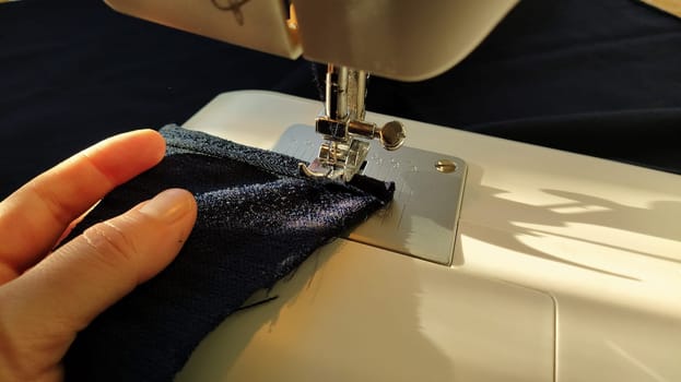 Sewing process on a modern sewing machine, close-up. Sewing machine foot with a needle. Dark blue fabric is trimmed with double wrapping of the fabric edge. Natural sunlight.