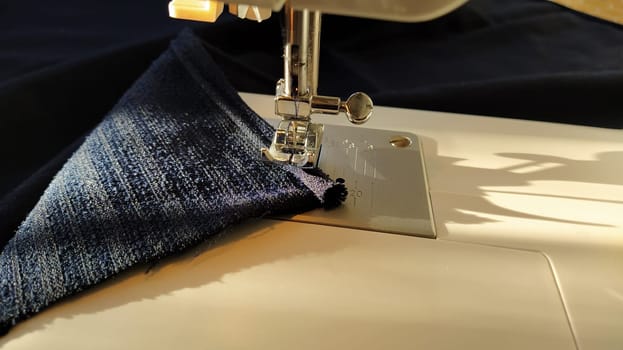 Sewing process on a modern sewing machine, close-up. Sewing machine foot with a needle. Dark blue fabric is trimmed with double wrapping of the fabric edge. Natural sunlight.