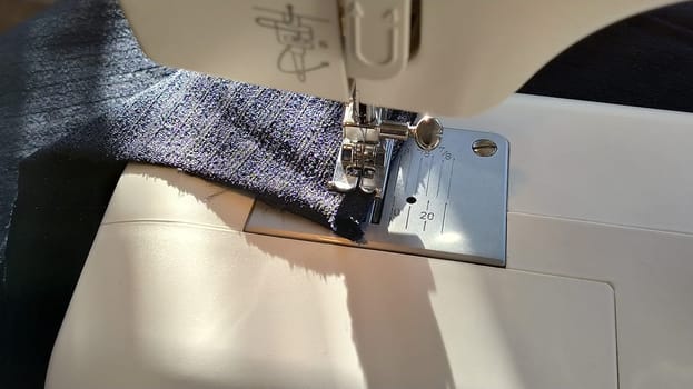 Sewing process on a modern sewing machine, close-up. Sewing machine foot with a needle. Dark blue fabric is trimmed with double wrapping of the fabric edge. Natural sunlight.