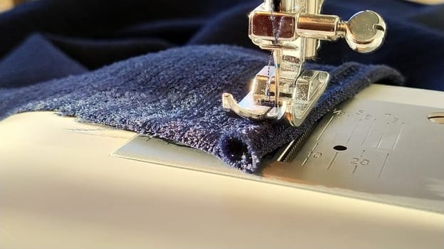 Sewing process on a modern sewing machine, close-up. Sewing machine foot with a needle. Dark blue fabric is trimmed with double wrapping of the fabric edge. Natural sunlight.