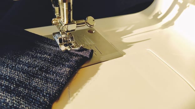 Sewing process on a modern sewing machine, close-up. Sewing machine foot with a needle. Dark blue fabric is trimmed with double wrapping of the fabric edge. Natural sunlight. Copy space.