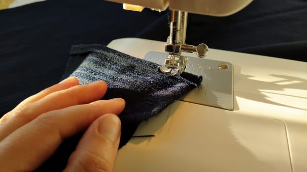 Sewing process on a modern sewing machine, close-up. Sewing machine foot with a needle. Dark blue fabric is trimmed with double wrapping of the fabric edge. Natural sunlight.