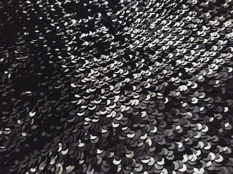 Sequin fabric. Black sequins with white and gray sheen. Reflection of light on individual elements in a chaotic manner. Sewing or Christmas decorative background.