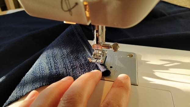 Sewing process on a modern sewing machine, close-up. Sewing machine foot with a needle. Dark blue fabric is trimmed with double wrapping of the fabric edge. Natural sunlight.