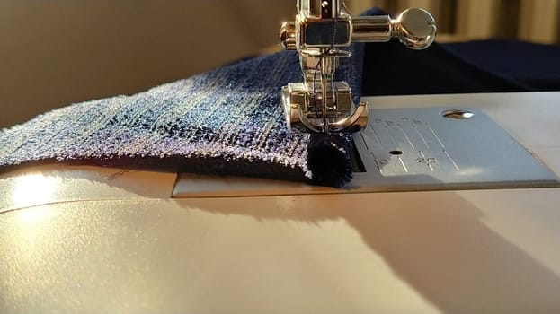 Sewing process on a modern sewing machine, close-up. Sewing machine foot with a needle. Dark blue fabric is trimmed with double wrapping of the fabric edge. Natural sunlight.