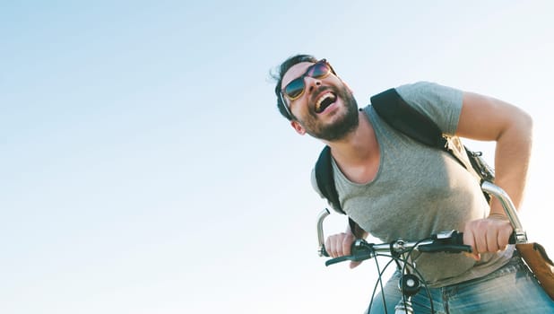 Enthusiastic hipster guy with retro sunglasses having outdoor fun and riding a bicycle. Active sport man with excited face expression exploring and traveling by mountain bike on the road. Vintage film filter style image.