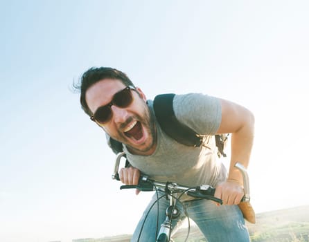 Enthusiastic hipster guy with retro sunglasses having outdoor fun and riding a bicycle. Active sport man with excited face expression exploring and traveling by mountain bike on the road. Vintage film filter style image.