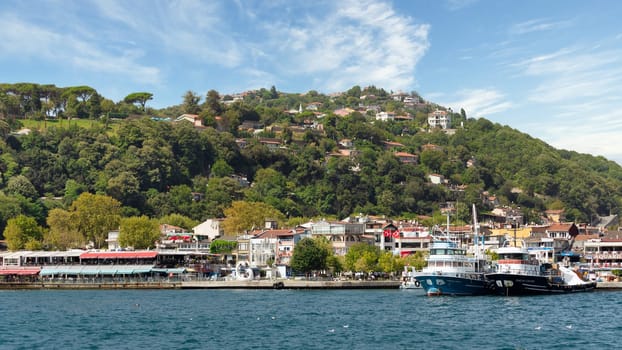 View from the sea of the green mountains of the Europian side of Bosphorus strait, with docked boats, traditional houses and dense trees in a summer day, Istanbul, Turkey