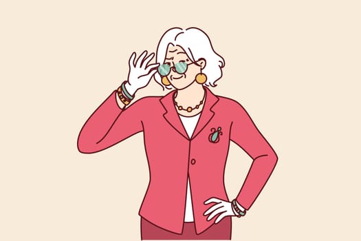 Elderly woman in elegant suit for going to work or dinner party adjusts glasses in front of eyes. Gray-haired grandmother of retirement age dressed in fashionable elegant clothes looks at camera