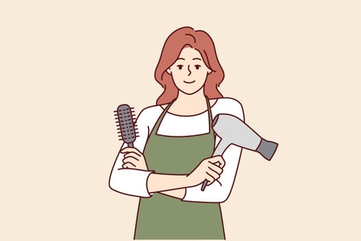 Woman professional hairdresser from barbershop holding comb and hair dryer to care for client hairstyle. Girl working as hairdresser in beauty salon stands with arms crossed and looks at camera
