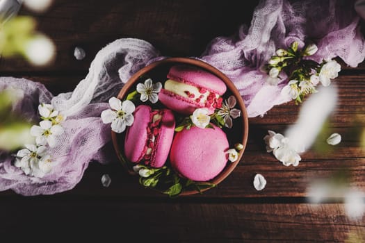 Top view on pink strawberry macaron cookies in ceramic bowl on wooden brown deck boards background, white apple blossom flowers. Colorful composition flat lay with sweet small French macaroon cakes