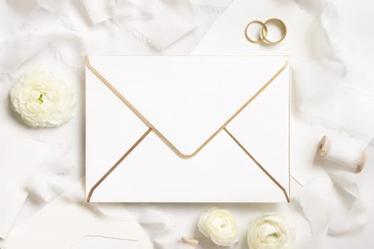 Blank envelope near cream roses, white silk ribbons and wedding rings top view, wedding mockup. Romantic scene with vertical card and pastel flowers flat lay. Valentines, Spring or Mothers day concept
