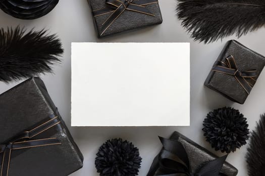 Blank card between Wrapped Gift Boxes, black paper flowers and feathers, mockup, copy space. Dark composition with horizontal paper card for Birthday, Anniversary or black party present