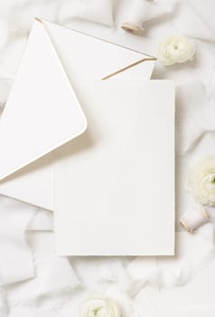 Blank card and envelope near cream roses and white silk ribbons top view, wedding mockup. Romantic scene with vertical card and pastel flowers flat lay. Valentines, Spring or Mothers day concept