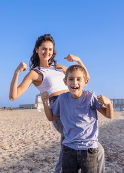 Happy Mother and son Showing strong arms on beach in a sunny day. Toddler kid boy spending time together with his Mom and having fun near the sea at sunset. Family together