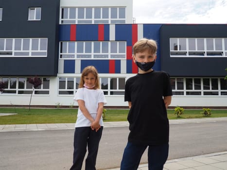 A blond boy 8 years old and a blond girl 7 years old are standing against the background of a modern school and looking at the camera. Children wear face masks. Black and white t-shirts.