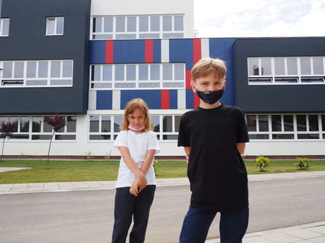 A blond boy 8 years old and a blond girl 7 years old are standing against the background of a modern school and looking at the camera. Children wear face masks. Black and white t-shirts.