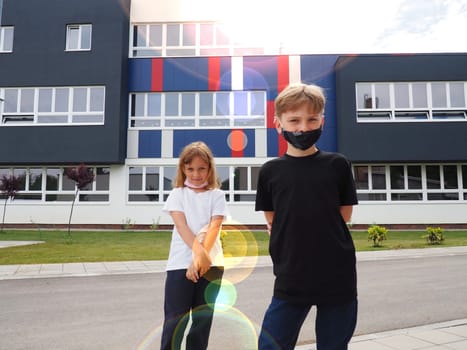 A blond boy 8 years old and a blond girl 7 years old are standing against the background of a modern school and looking at the camera. Children wear face masks. Sun glare.