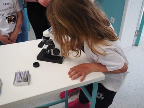 Sremska Mitrovica, Serbia, May 15, 2021. A girl of 7 years old in a white T-shirt looks with one eye through a microscope. Biology lesson. The process of teaching children. School lessons.