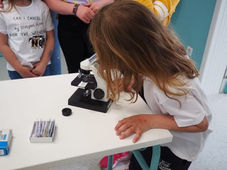 Sremska Mitrovica, Serbia, May 15, 2021. A girl of 7 years old in a white T-shirt looks with one eye through a microscope. Biology lesson. The process of teaching children. School lessons.