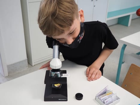 Sremska Mitrovica, Serbia, May 15, 2021. A boy of 8 years old in a black T-shirt looks with one eye through a microscope. Biology lesson. The process of teaching children. School lessons.