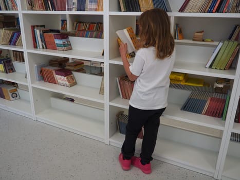 Sremska Mitrovica, Serbia May 15, 2021. A 7-year-old girl in a white T-shirt stands near the bookshelf in the library and reads a book. School education. Love for literature and reading.