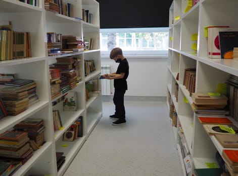 Sremska Mitrovica, Serbia May 15, 2021 An 8-year-old boy wearing a mask and a black T-shirt stands near the bookshelf in the library and reads a book. School education. Love for literature and reading.
