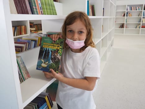 Sremska Mitrovica, Serbia May 15, 2021 A 7-year-old girl wearing a mask and a white T-shirt stands near a bookshelf in a library and touches books with hands. School education, literature and reading.
