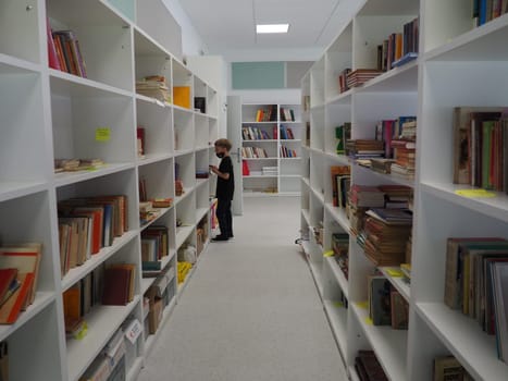 Sremska Mitrovica, Serbia May 15 2021 An 8-year-old boy wearing a mask and a black T-shirt stands near the bookshelf in the library and reads a book. School education. Love for literature and reading.