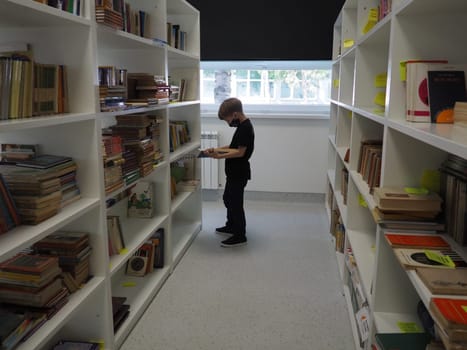 Sremska Mitrovica, Serbia May 15 2021 An 8-year-old boy wearing a mask and a black T-shirt stands near the bookshelf in the library and reads a book. School education. Love for literature and reading