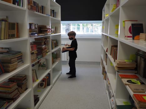 Sremska Mitrovica, Serbia May 15 2021 An 8-year-old boy wearing a mask and a black T-shirt stands near the bookshelf in the library and reads a book. School education. Love for literature and reading