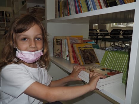 Sremska Mitrovica, Serbia May 15, 2021 A 7-year-old girl wearing a mask and a white T-shirt stands near a bookshelf in a library and touches books with hands. School education, literature and reading.