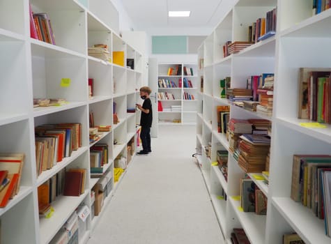 Sremska Mitrovica, Serbia May 15 2021 An 8-year-old boy wearing a mask and a black T-shirt stands near the bookshelf in the library and reads a book. School education. Love for literature and reading.
