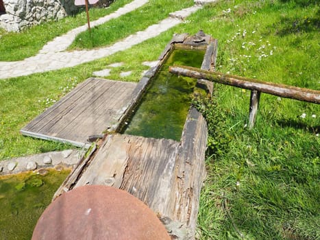Wooden water trough or livestock drinker. A hewn or hollowed-out log used in rural life. Hollow wooden gutter for water supply. Metal cover of the well and wooden walkways. Green grass on the lawn.