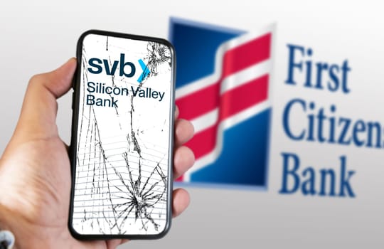 New York, US, March 2023: Hand holding a phone with Silicon Valley Bank logo on cracked screen. First Citizens Bank logo blurred on white background. Illustrative editorial