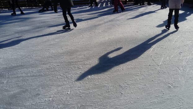 Children ride in a city park on an ice rink. Feet skater while skating on ice. The low winter sun weakly illuminates the ice. Dark shapes and long shadows on the surface. Sports movements.