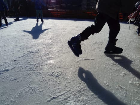 Children ride in a city park on an ice rink. Feet skater while skating on ice. The low winter sun weakly illuminates the ice. Dark shapes and long shadows on the surface. Sports movements.