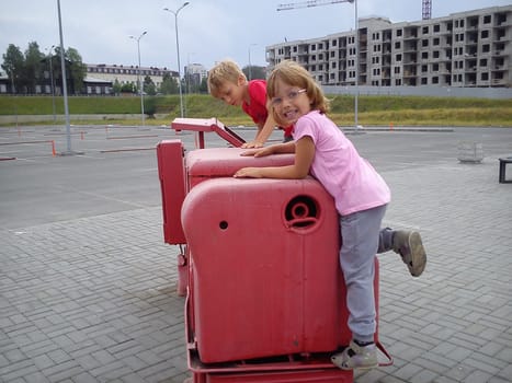 Children climb on the old factory machine, exposed to the street. Factory tractor-machine painted pink paint. Girl laughing boy is serious. Children with blond hair. Construction in the background.