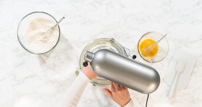 Top view of an electric mixer and bowls containing ingredients for Classic Sugar Cookie Dough. The mixer is placed over a bowl with partially mixed wet ingredients, while nearby bowls hold flour and a beaten egg mixture on a light marble surface.