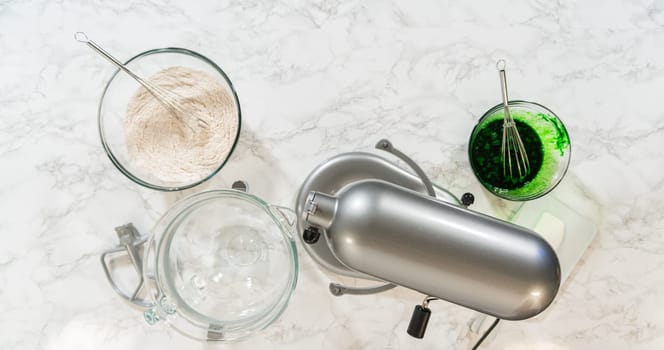 Ingredients organized around a stand mixer, ready to be combined for Vibrant Green Gingerbread Cookies for Festive Baking. Green color mixture and dry ingredients are set beside butter and sugar in the mixer bowl.