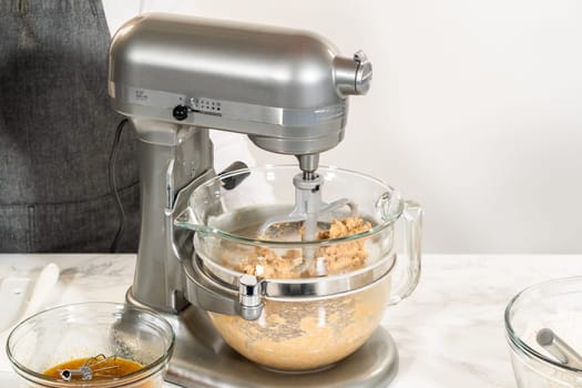 Side view of a stand mixer with ingredients for the Amber Gingerbread Cookie Dough in the mixing bowl, positioned on a marble countertop with a separate bowl of wet ingredients nearby, capturing the preparation stage of dough mixing.