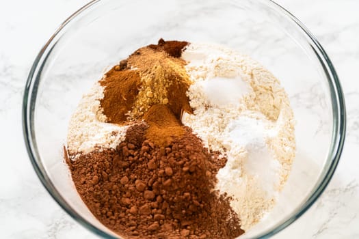 Mixing dry ingredients for Chocolate Gingerbread Cookie Dough, including flour, cocoa powder, cinnamon, ground ginger, and cloves in a glass bowl. The ingredients are layered and ready to be combined, creating a rich, aromatic base for the cookie dough.