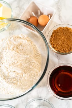 Side view of various baking ingredients arranged around a large glass bowl filled with flour on a marble surface. Ingredients include butter, eggs, brown sugar, spices, and maple syrup, showcasing the setup for Amber Gingerbread Cookie Dough.
