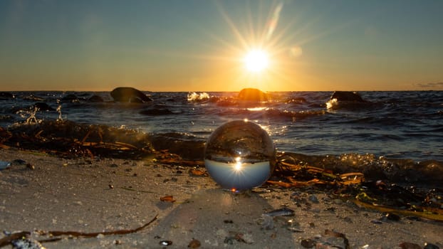 A glass ball lies in the sand in front of the waves, beach with algae and the sea and the setting sun are reflected in the ball