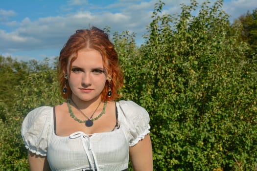 Portrait of a pretty young woman wearing white top, standing outdoors with green trees and blue sky