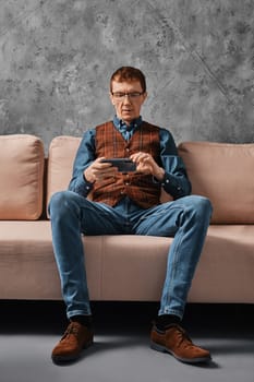 Serious middle aged man sitting on sofa and watching video on smartphone