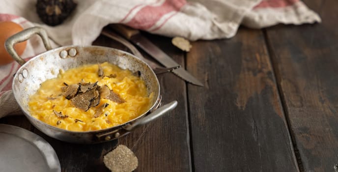 Scrambled eggs with sliced fresh black truffles from Italy served in a a frying pan near fork and knife on wood, close up, copy space. Italian cousine, gourmet breakfast