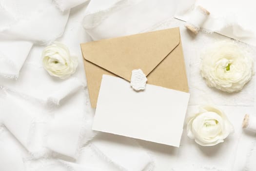 Blank card and envelope near cream roses and white silk ribbons top view, wedding mockup. Romantic scene with vertical card and pastel flowers flat lay. Valentines, Spring or Mothers day concept