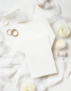 Blank card and envelope near cream roses, white silk ribbons and wedding rings top view, wedding mockup. Romantic scene with vertical card and pastel flowers flat lay. Valentines, Spring or Mothers day concept