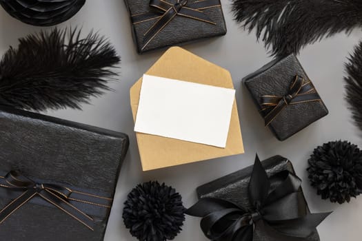Blank card and envelope between Wrapped Gift Boxes, black paper flowers and feathers, mockup, copy space. Dark composition with horizontal paper card for Birthday, Anniversary or black party present
